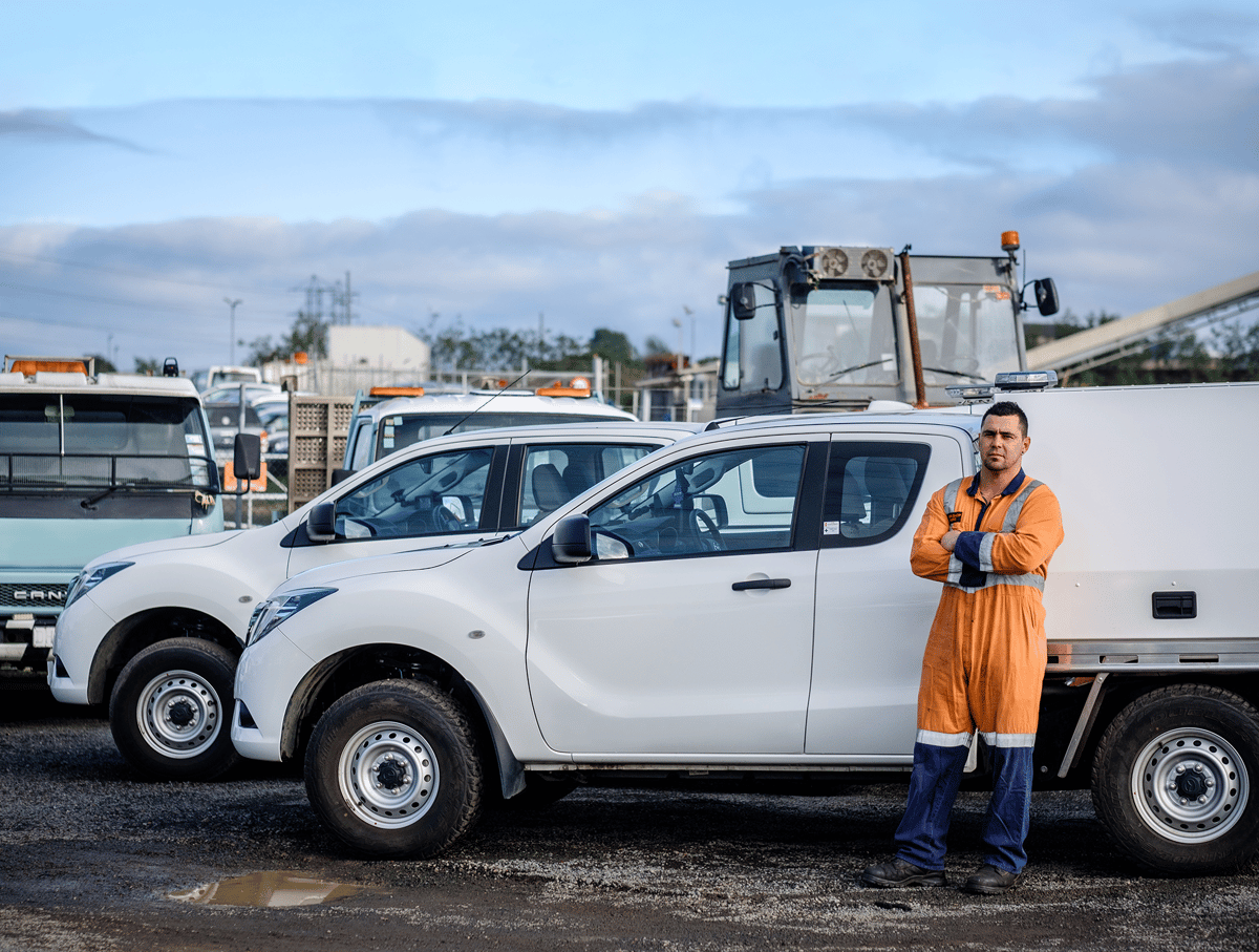 Worker in orange hi-vis overalls standing beside white utes in a fleet yard
