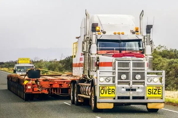 Truck with oversize signs towing a low-loader trailer on a highway, escort vehicle behind