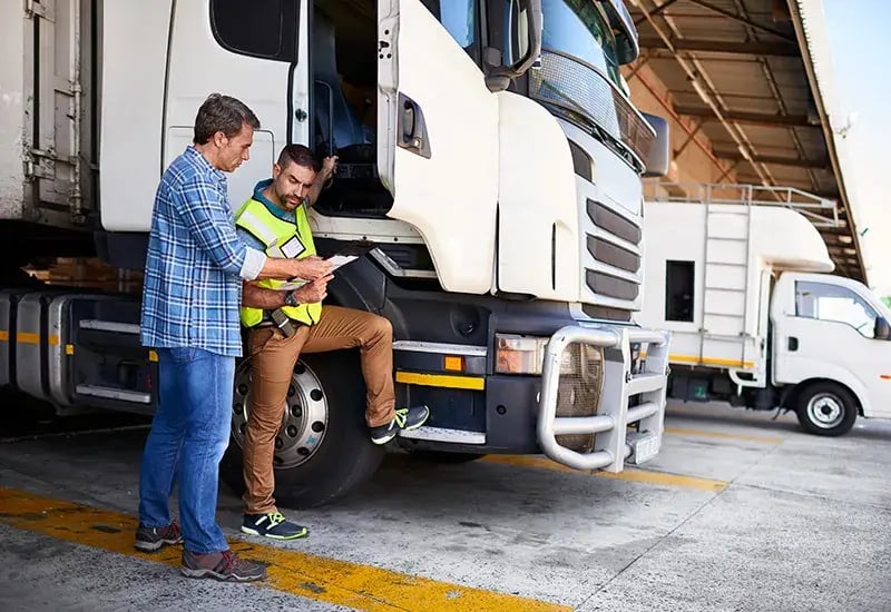 Two people reviewing a document beside a truck, one in a hi-vis vest sitting on the cab step