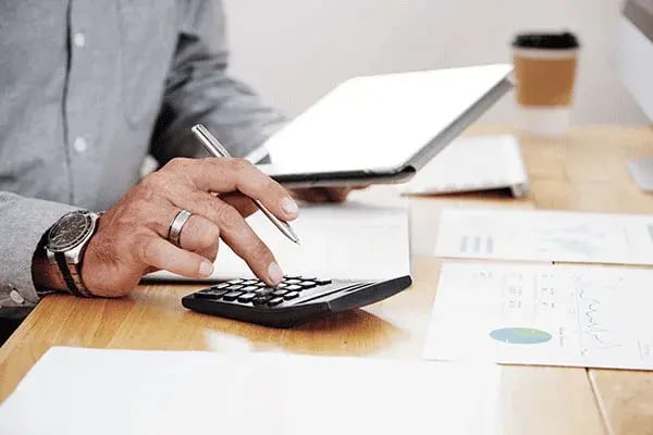 Person working out figures on a calculator beside a tablet and printed charts