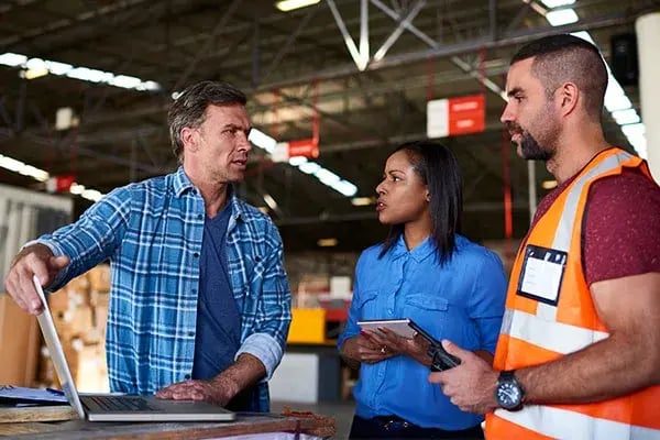 Three workers talking in a warehouse, one with a laptop and one with a tablet