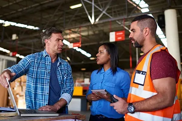 Three people talking in a warehouse, one leaning on a laptop and one holding a tablet