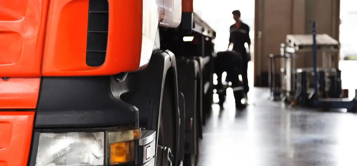 Front of an orange truck in a workshop, with workers beside its wheels in the background