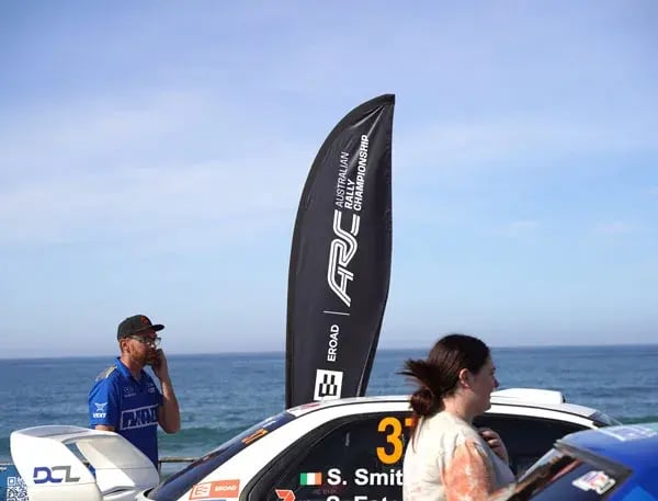 EROAD Australian Rally Championship flag by the sea, beside a rally car and people at the event