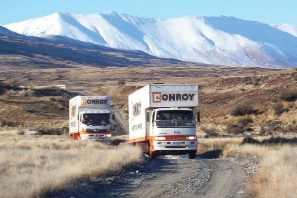 Two Conroy removal trucks on a gravel road with snow-covered mountains behind