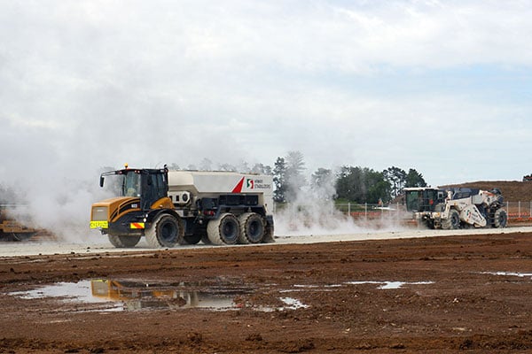 Hiway spreader truck and a road stabiliser working a muddy earthworks site in a cloud of white dust