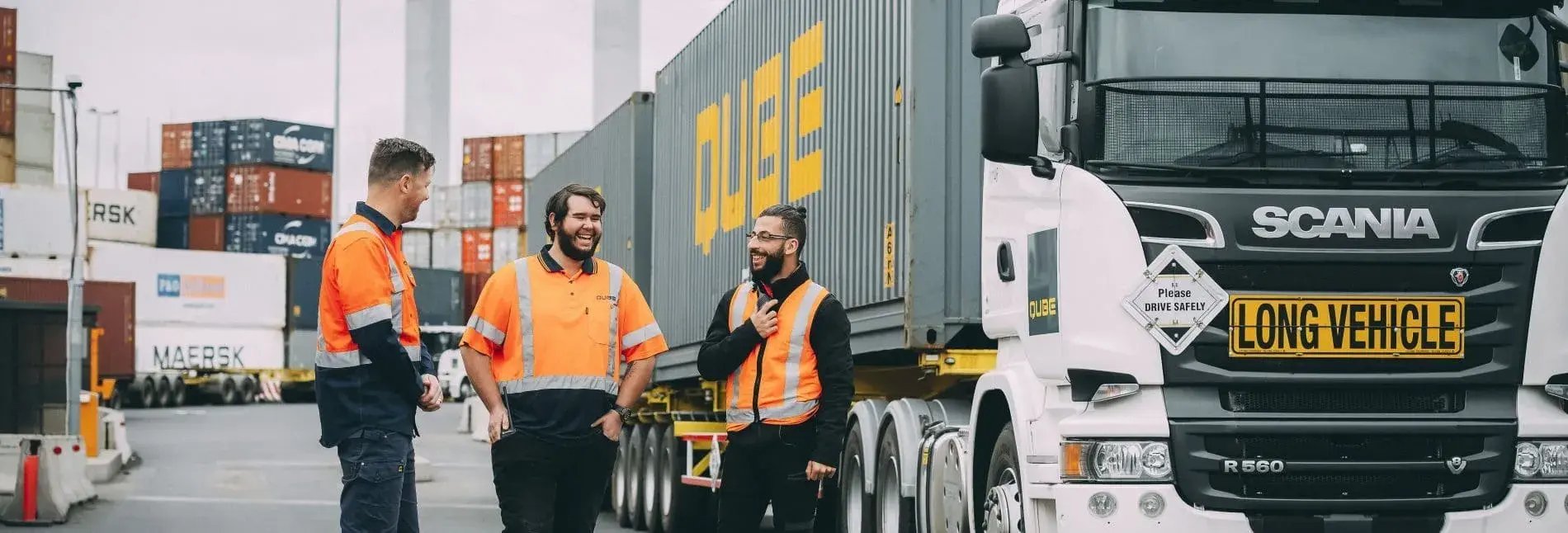 Three Qube workers laughing beside a Scania prime mover in a container yard