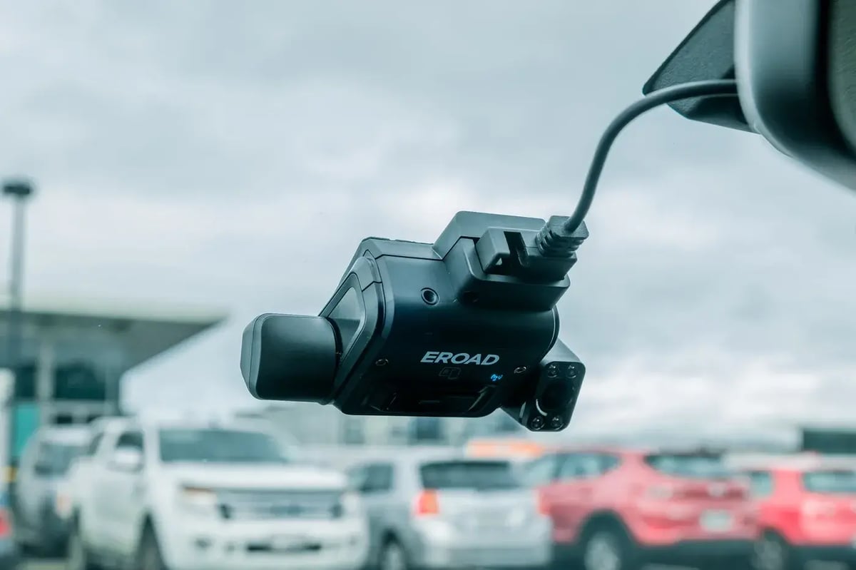 An EROAD Clarity dashcam mounted on a light vehicle windscreen, with its driver-facing camera and a car park beyond