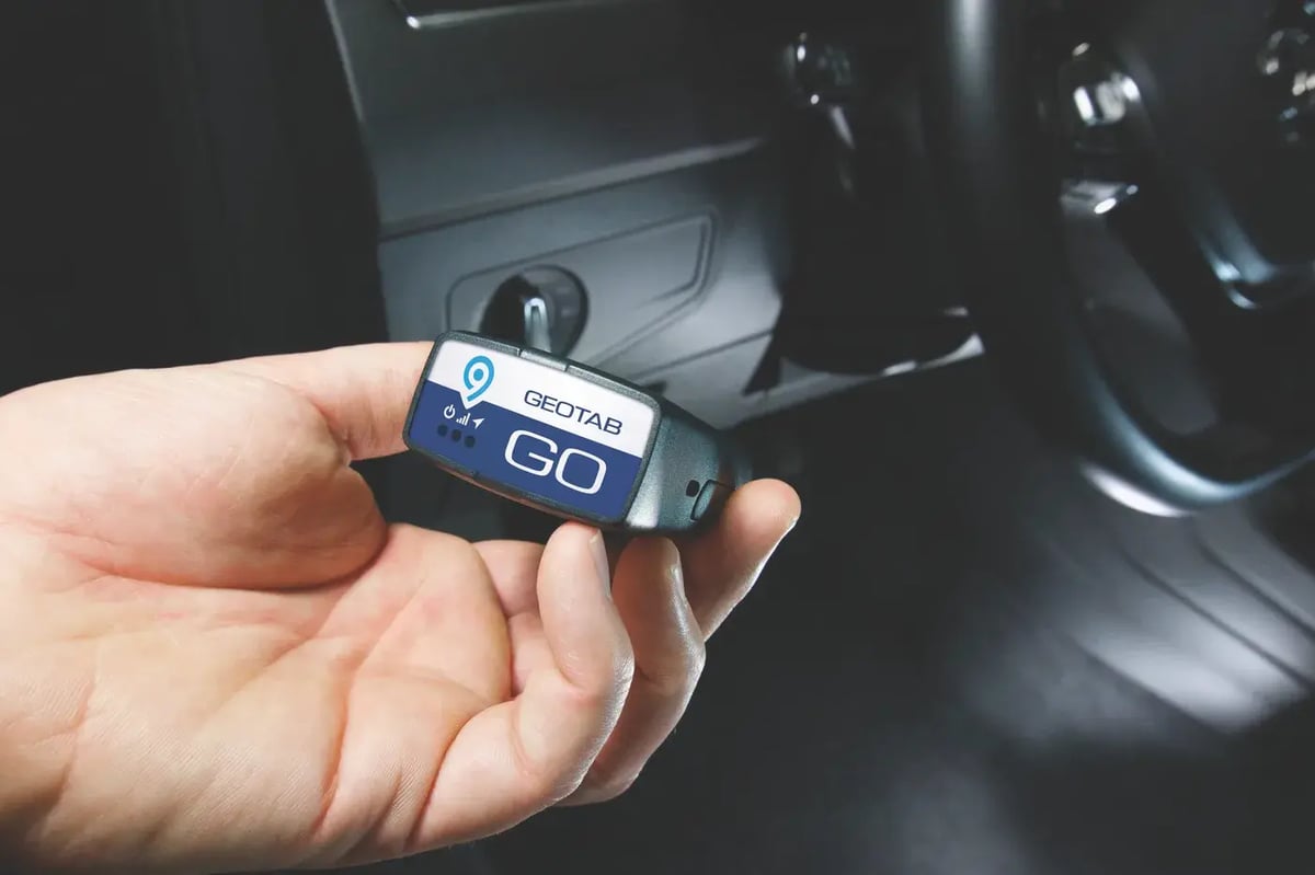 A hand holding a Geotab GO device next to a vehicle dashboard before plugging it into the OBD port
