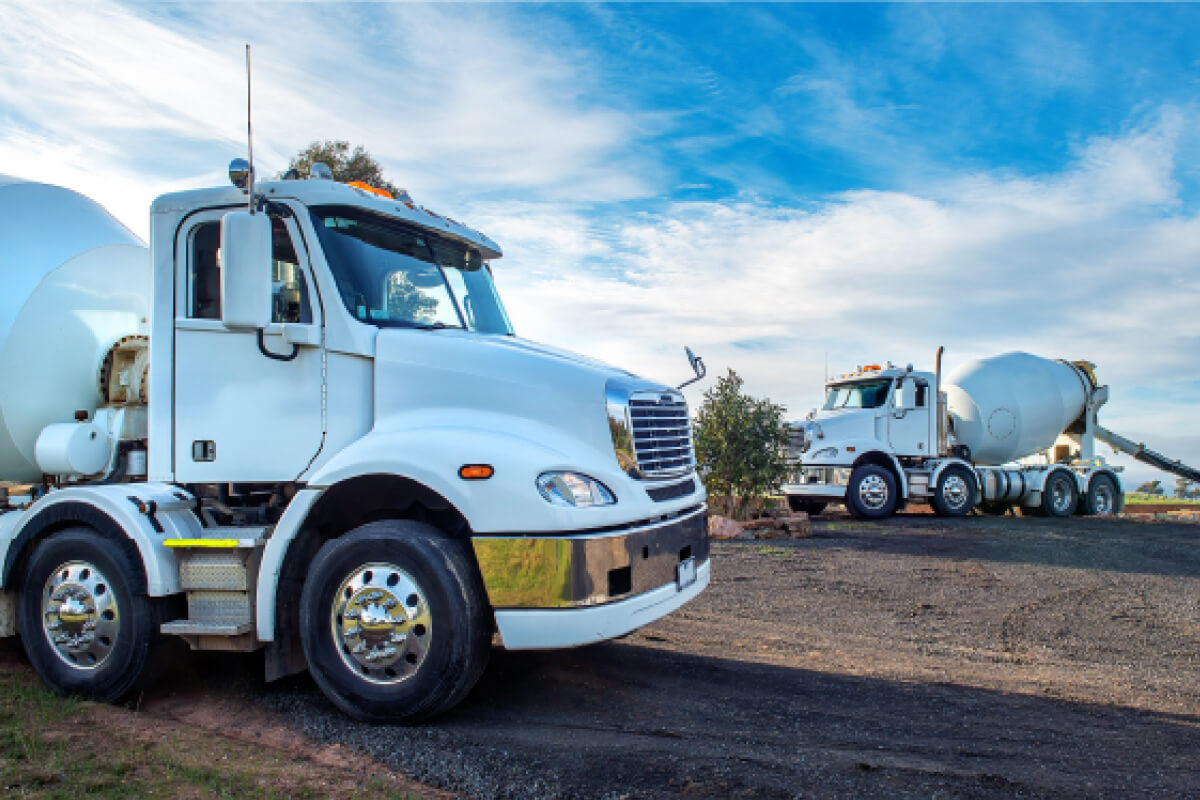 Two white concrete mixer trucks parked on a gravel yard under a blue sky