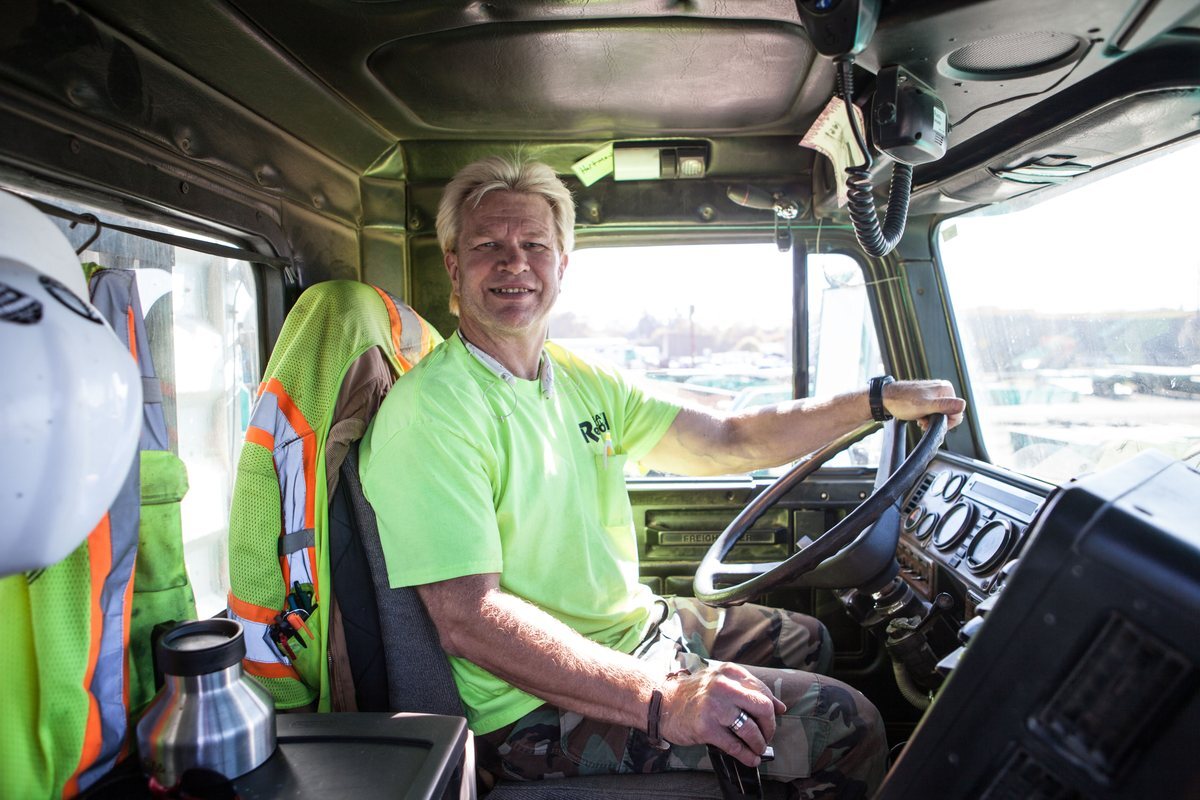 A truck driver at the wheel, with a hard hat and high visibility vest hanging in the cab