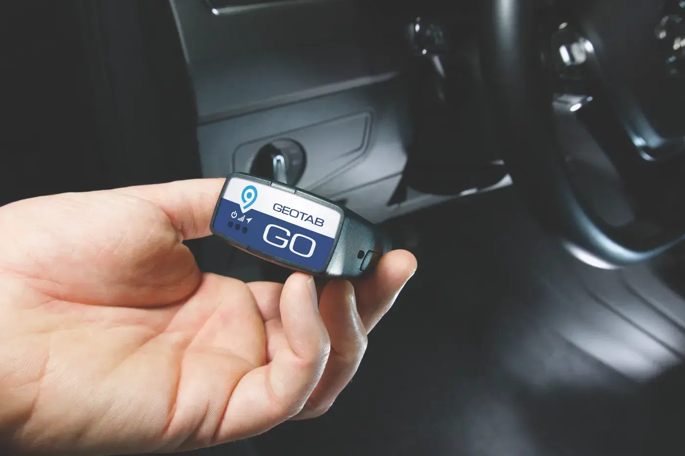 A hand holding a Geotab GO device next to a vehicle dashboard before plugging it into the OBD port